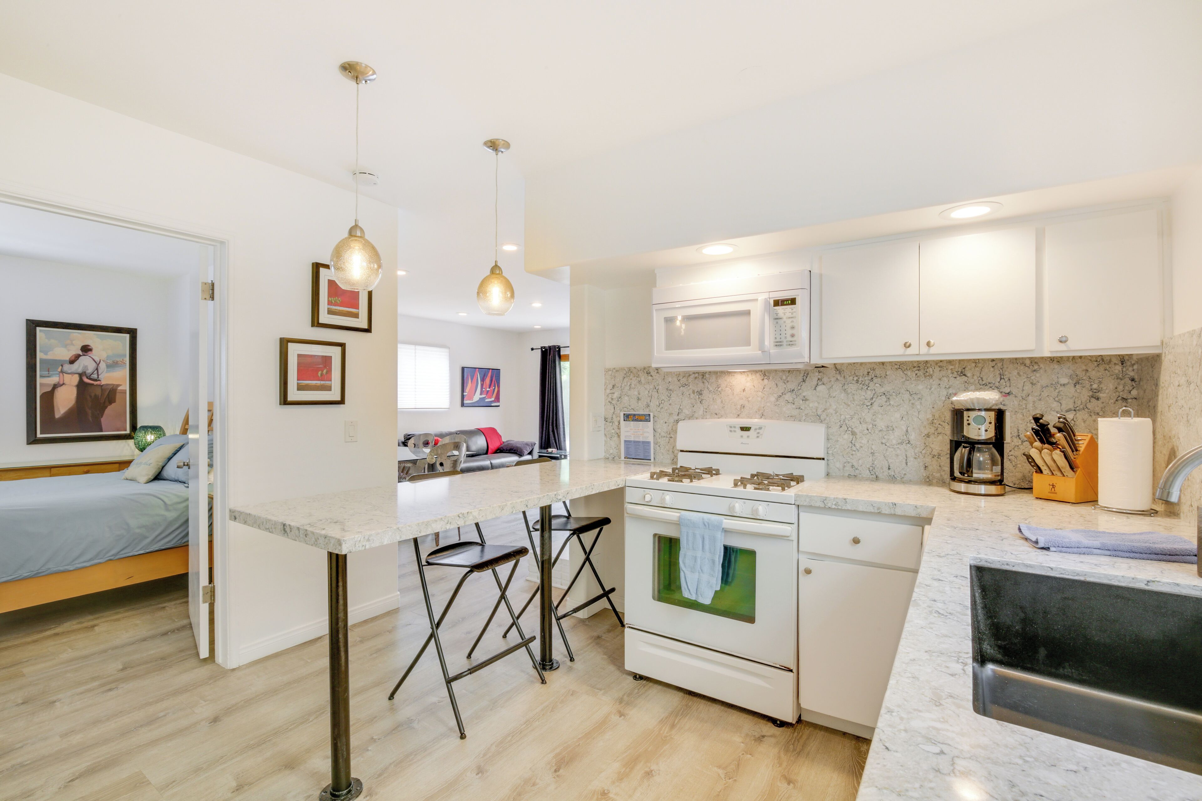 A modern kitchen with white cabinets, a built-in microwave, and a gas stove. There is a bar area with stools and a painting on the wall.