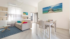 Ein modernes Wohnzimmer mit einer Couch, einem Glastisch und einem Essbereich mit Stühlen und einem Tisch. An der Wand hängt ein Gemälde einer Strandansicht.