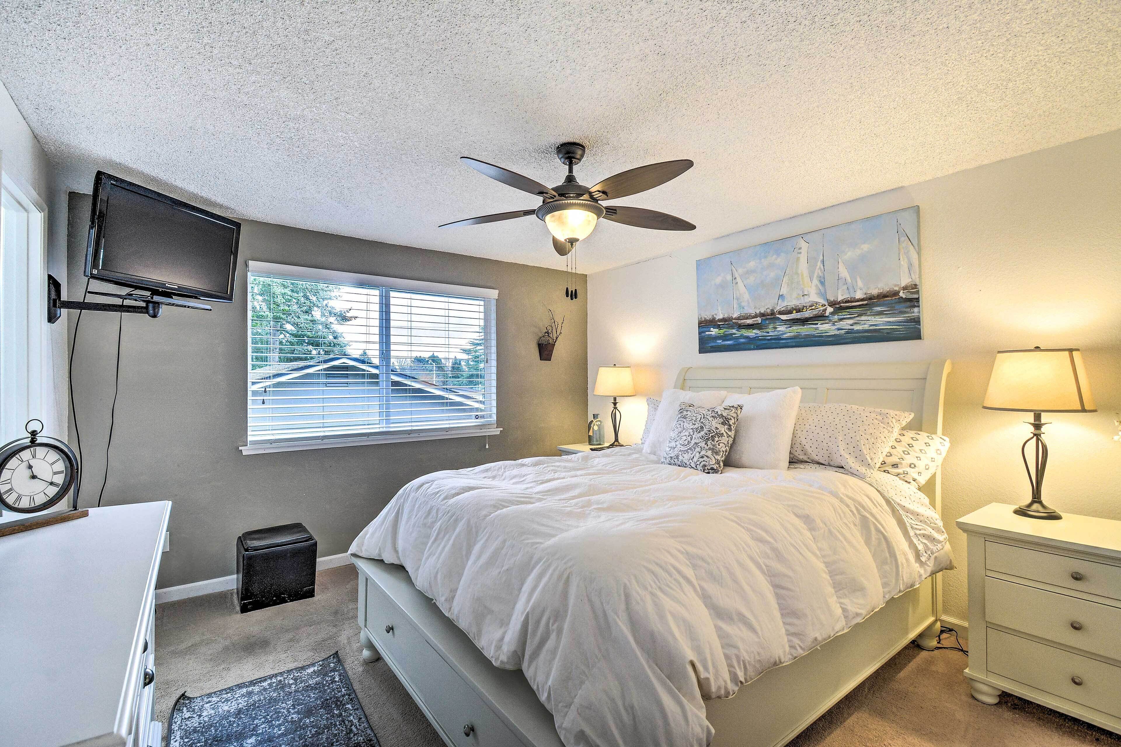 A bedroom with a bed, ceiling fan, TV, bedside lamps, a painting, and a window with blinds.