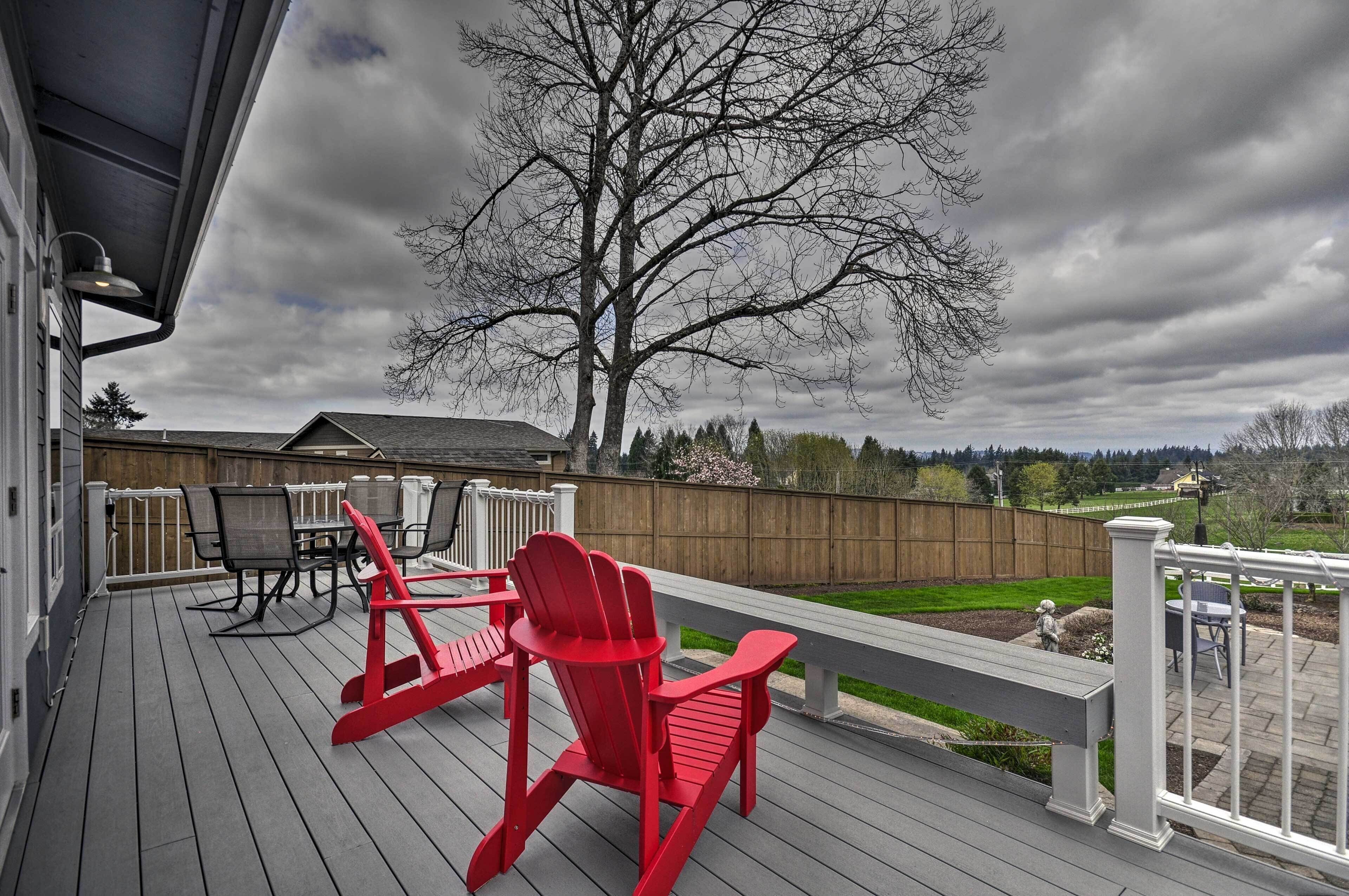 A wooden deck with red Adirondack chairs and a table, overlooking a backyard with a large tree and a wooden fence.
