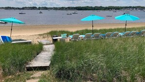 Ein Strand mit Liegestühlen und Sonnenschirmen, eine Holzpromenade, die zum Strand führt, und Boote auf dem Wasser.