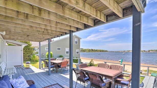 Eine überdachte Terrasse mit Gartenmöbeln, Blick auf einen See und ein Haus im Hintergrund.