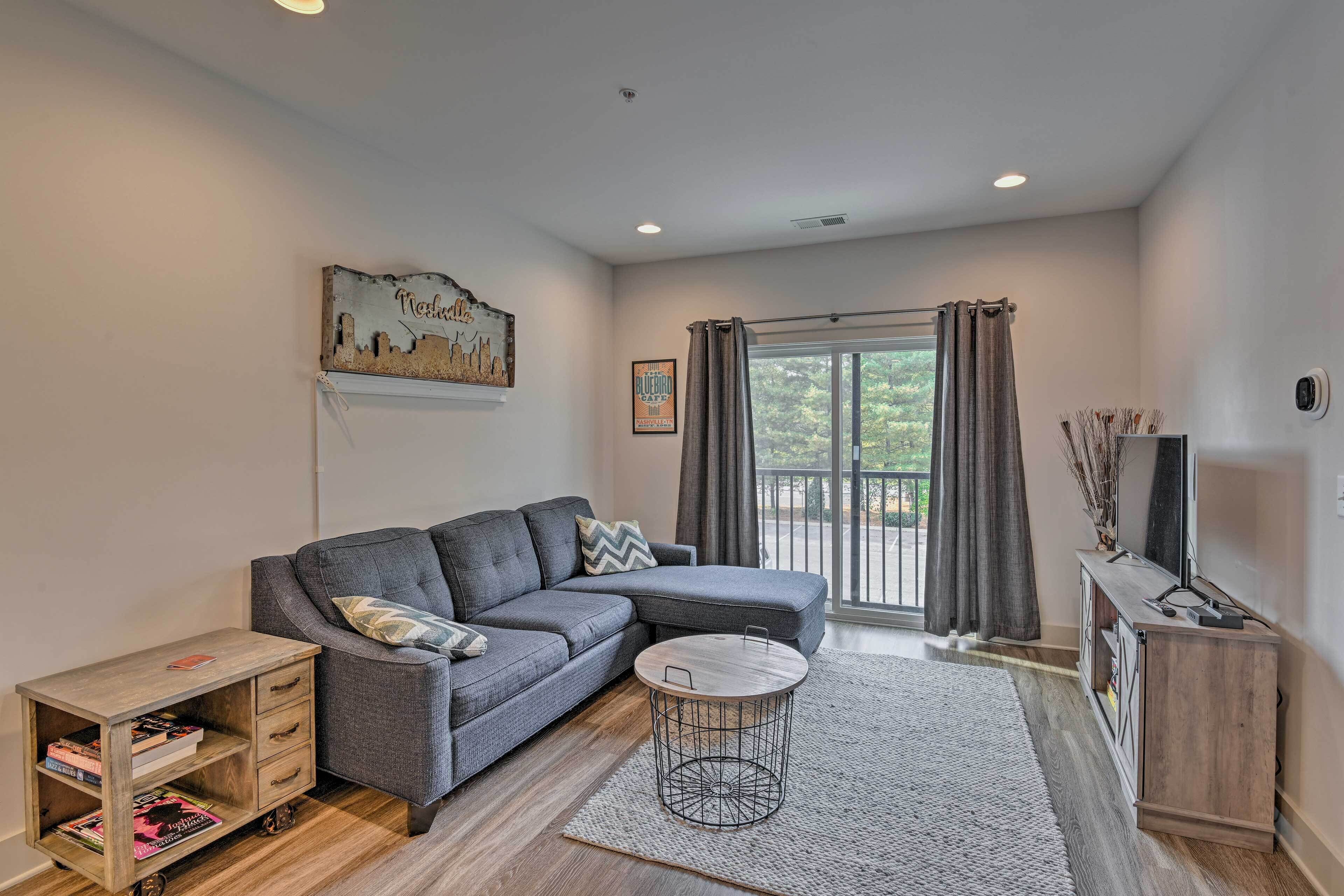 A modern living room with a grey sectional sofa, a small wooden side table with books, a round coffee table, and a large window with curtains.