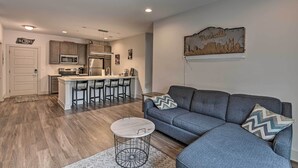 A modern living room with a gray sectional sofa, a round coffee table, and a kitchen area with wooden cabinets and stainless steel appliances. - Modern Nashville Condo: 7 Mi to Downtown! (Nashville)