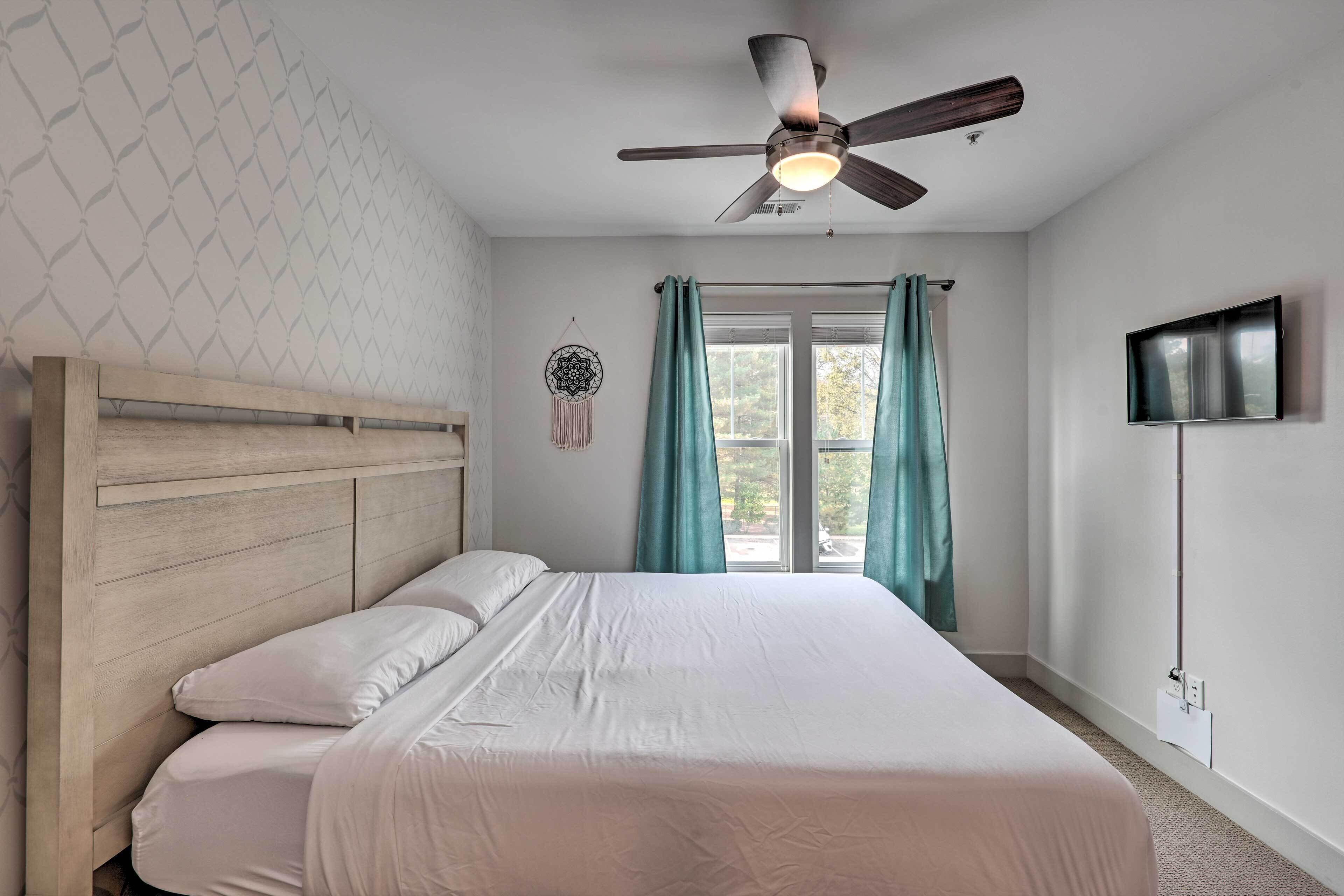 A bedroom with a wooden headboard, a bed with white bedding, teal curtains, a ceiling fan, a wall-mounted TV, and a dreamcatcher decoration.