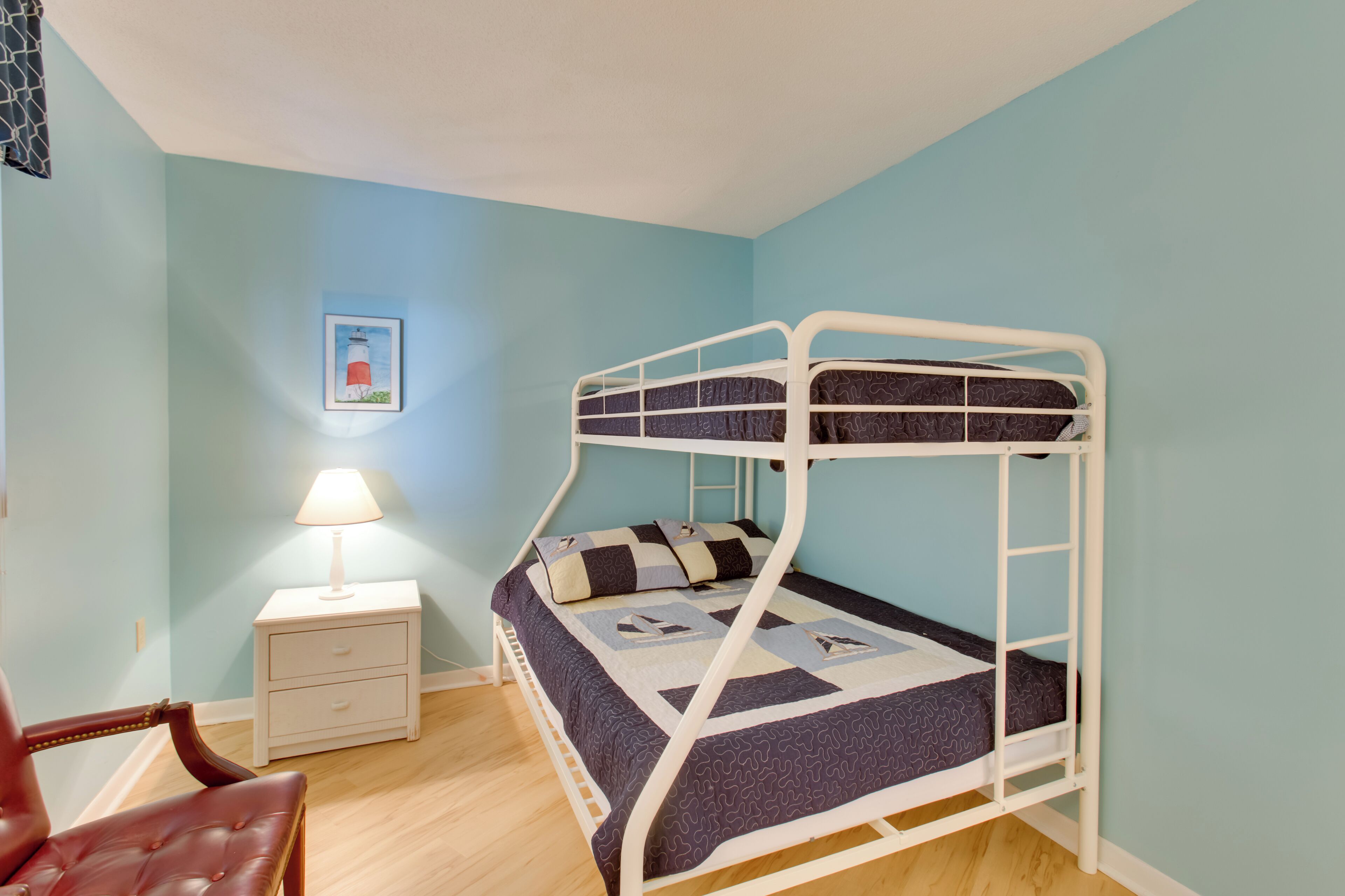 Una cama litera en una habitación con paredes azul claro, suelo de madera, una mesita de noche con lámpara y un cuadro en la pared.