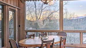 Une table à manger en bois, des chaises, un lustre et une grande fenêtre donnant sur un paysage de arbres et de montagnes.