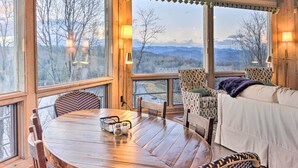 Une pièce chaleureuse avec une table à manger en bois, des chaises et un canapé. De grandes fenêtres offrent une vue sur les montagnes et les arbres.