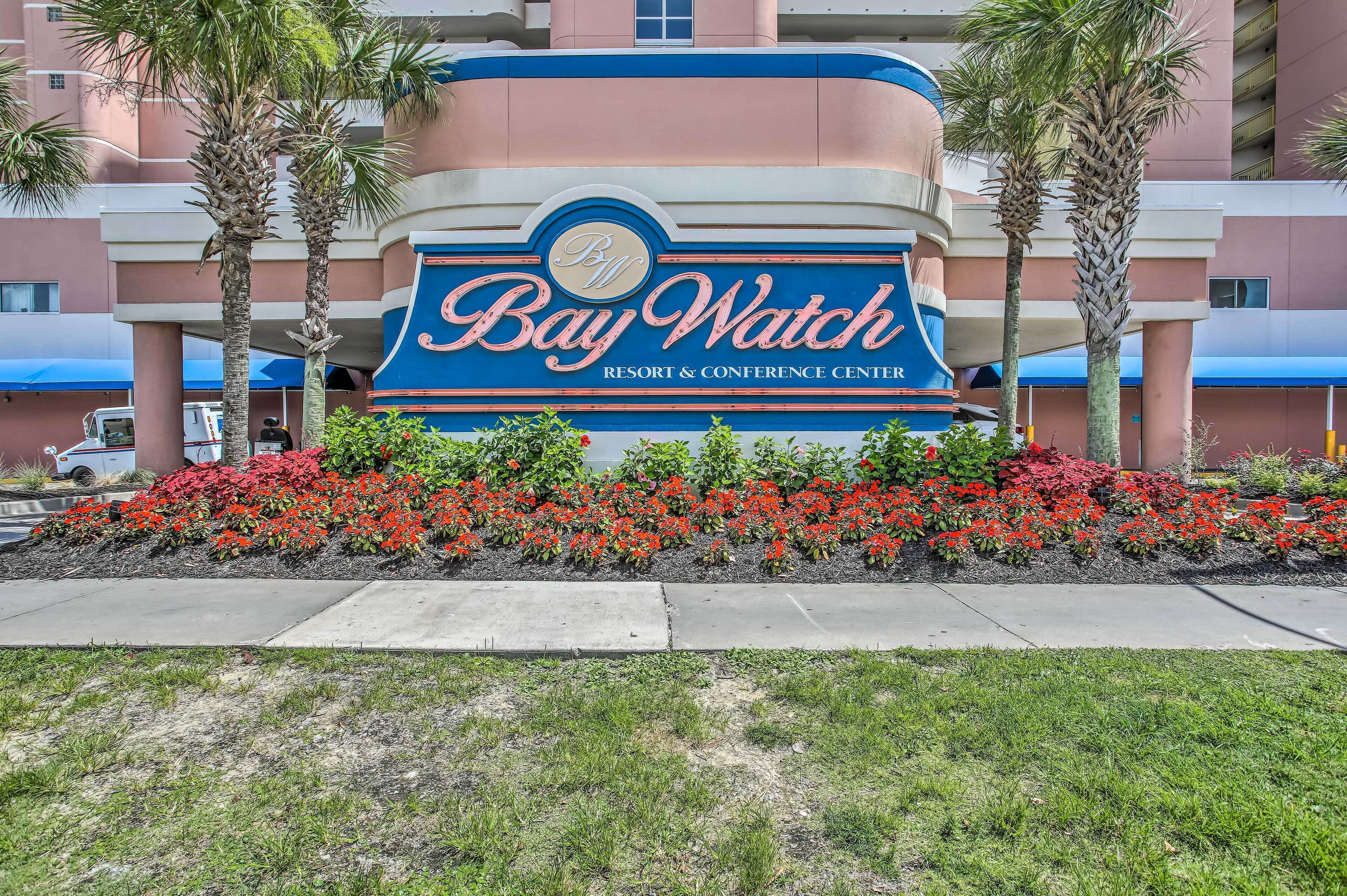 Une enseigne pour le Bay Watch Resort & Conference Center, avec un palmier et des fleurs rouges au premier plan.