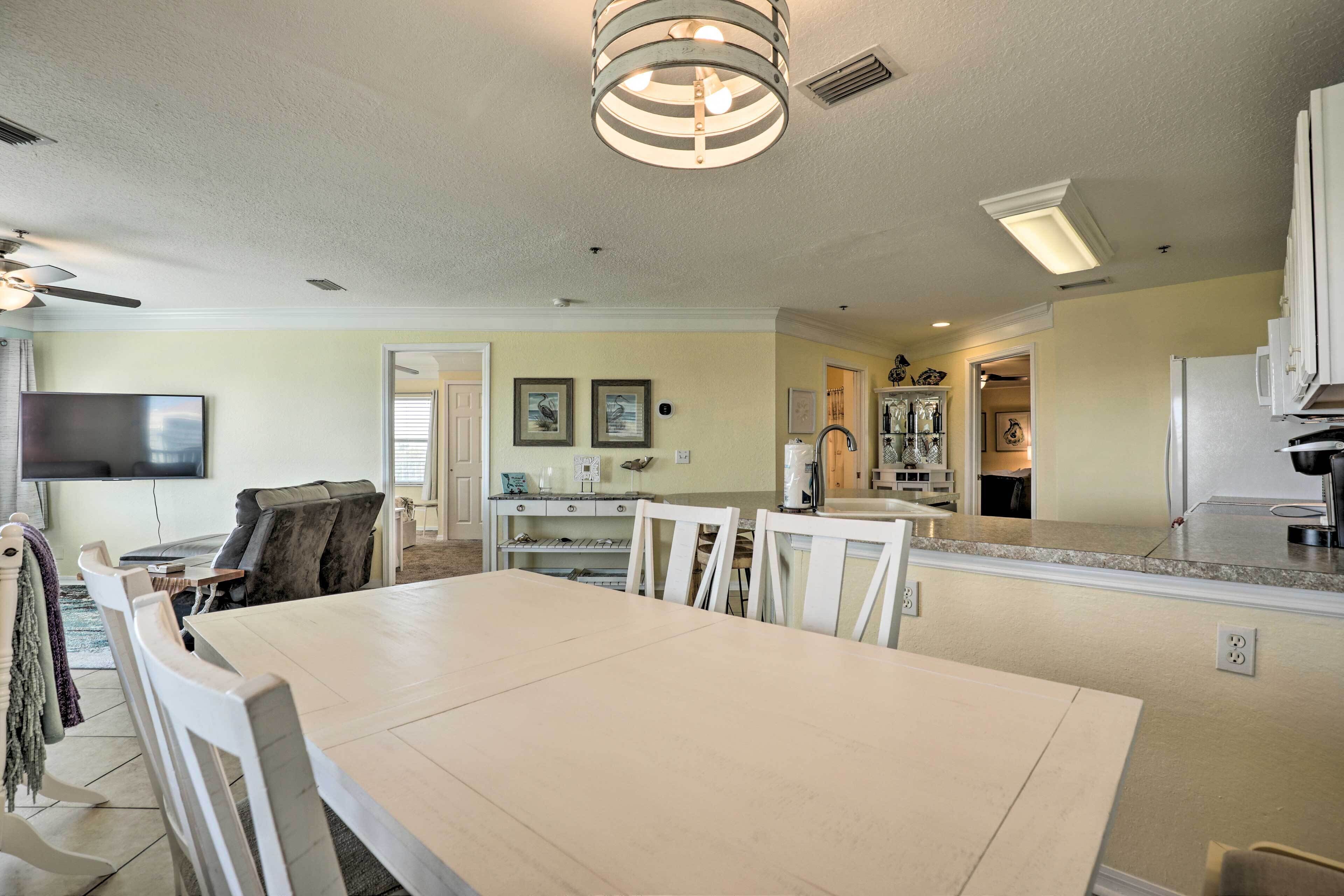A spacious living room with a large white dining table, a ceiling fan, and a flat-screen TV mounted on the wall.