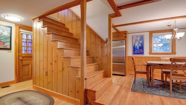 Una escalera de madera que conduce a un segundo piso, un comedor con mesa y sillas, y un refrigerador al fondo.