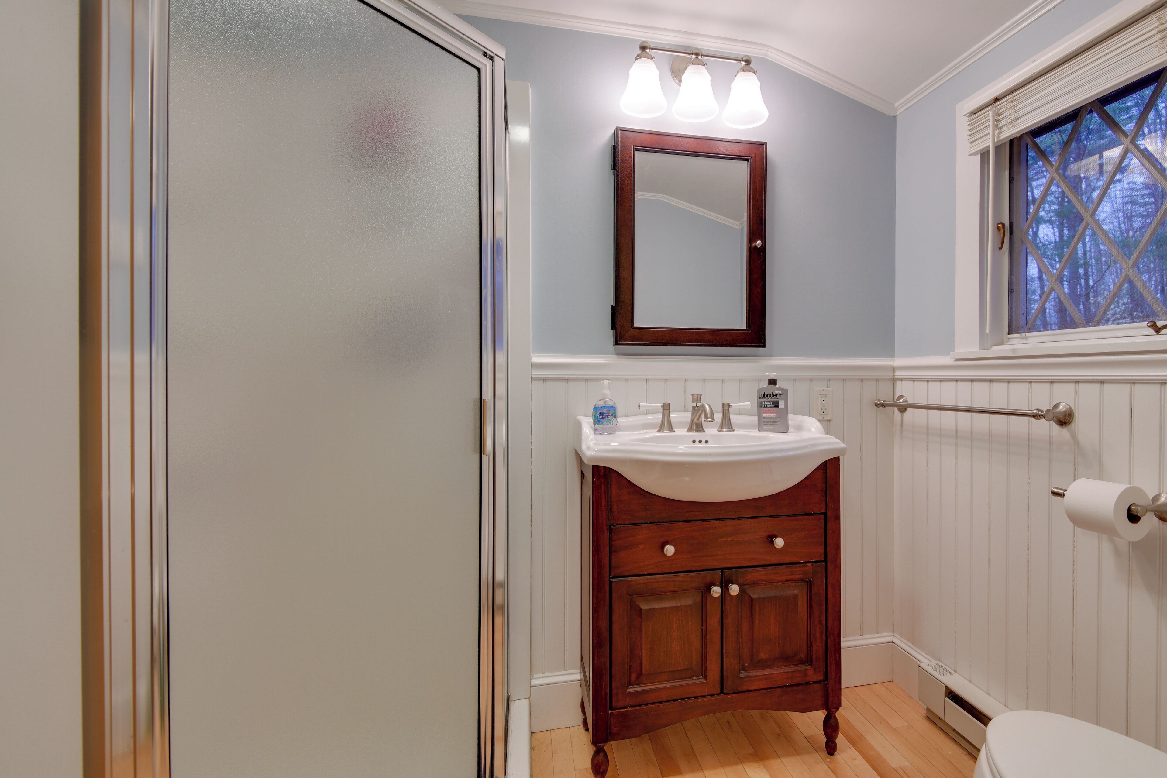 A bathroom with a wooden vanity, a white sink, and a mirror above it. There is a window with a cross pattern and a towel rack on the wall.