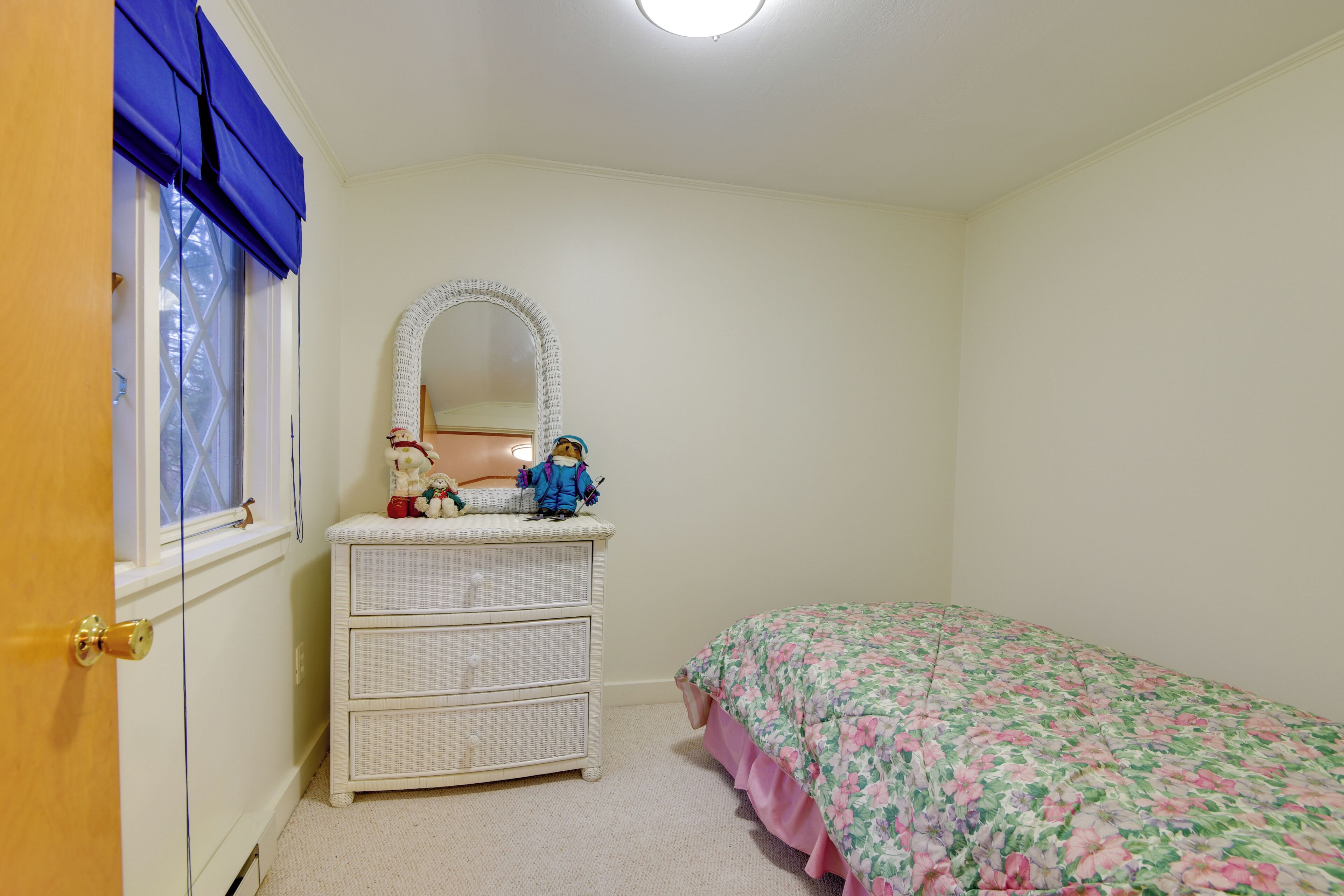 A bedroom with a white dresser, a mirror, a floral bedspread, and a window with blue blinds.