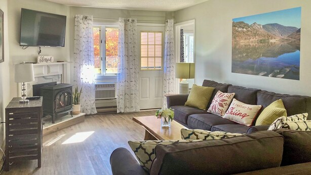 A cozy living room with a fireplace, a sofa with patterned cushions, and a large window with curtains. - Hike, Ski, Explore: Lincoln Condo w/ Pool Access (Lincoln)