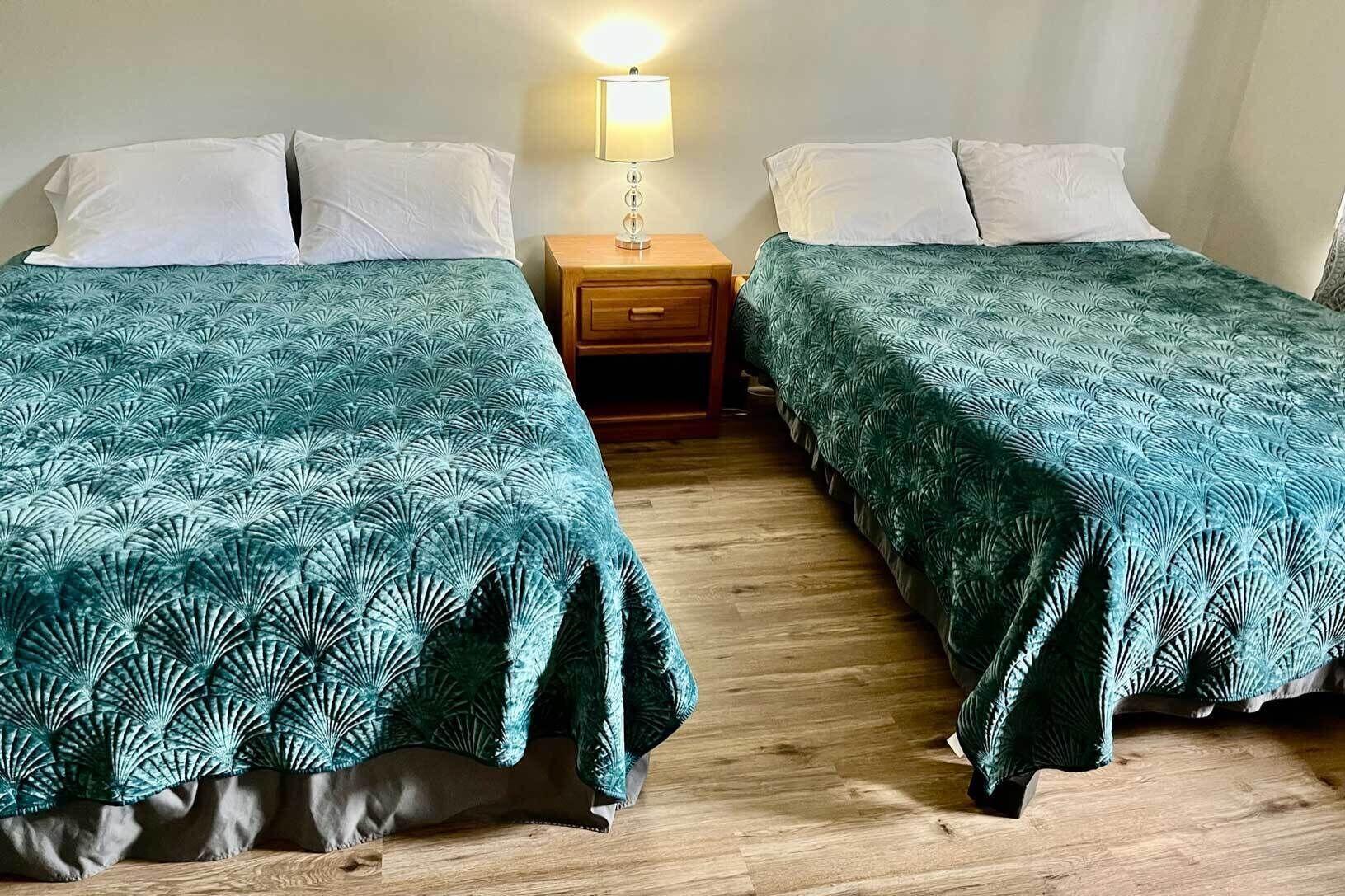 Deux lits simples avec des couvre-lits verts à motifs assortis, une table de chevet en bois avec une lampe et un plancher en bois.