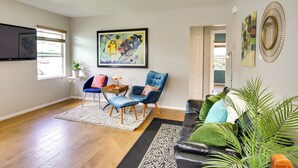 A living room with hardwood flooring, a blue armchair, a black sofa, and a coffee table. There are two paintings on the wall and a potted plant.