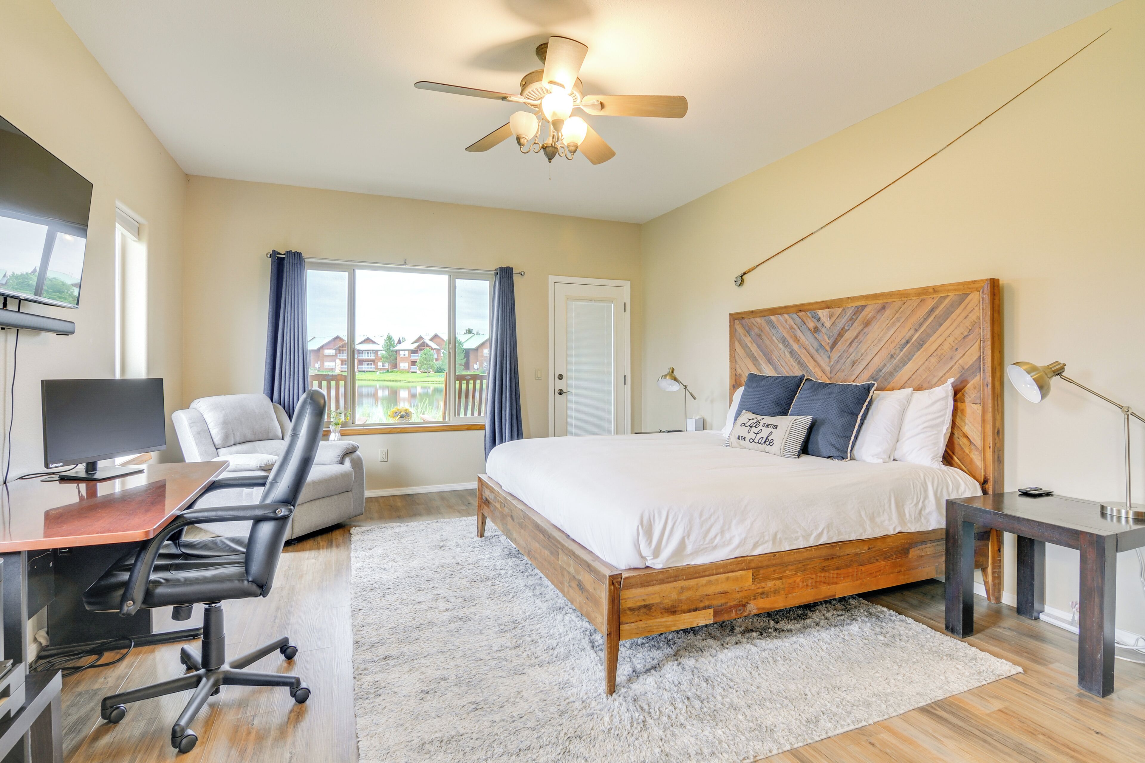 A bedroom with a bed, desk, chair, ceiling fan, and large windows.