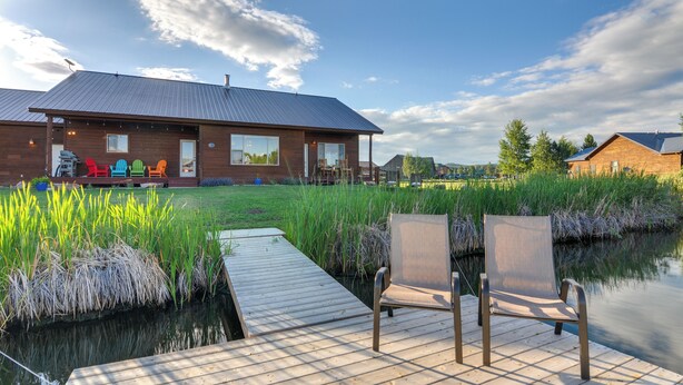 Ein Holzhaus mit einer Terrasse, die über einen Teich hinausreicht, zwei Stühle und ein Grill.