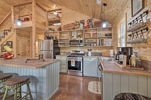 A rustic kitchen with wooden beams, a central island with stools, a refrigerator, stove, microwave, and various kitchen utensils on shelves. - Peaceful Family Cabin on 10 Acres w/ Game Room! (Heflin)
