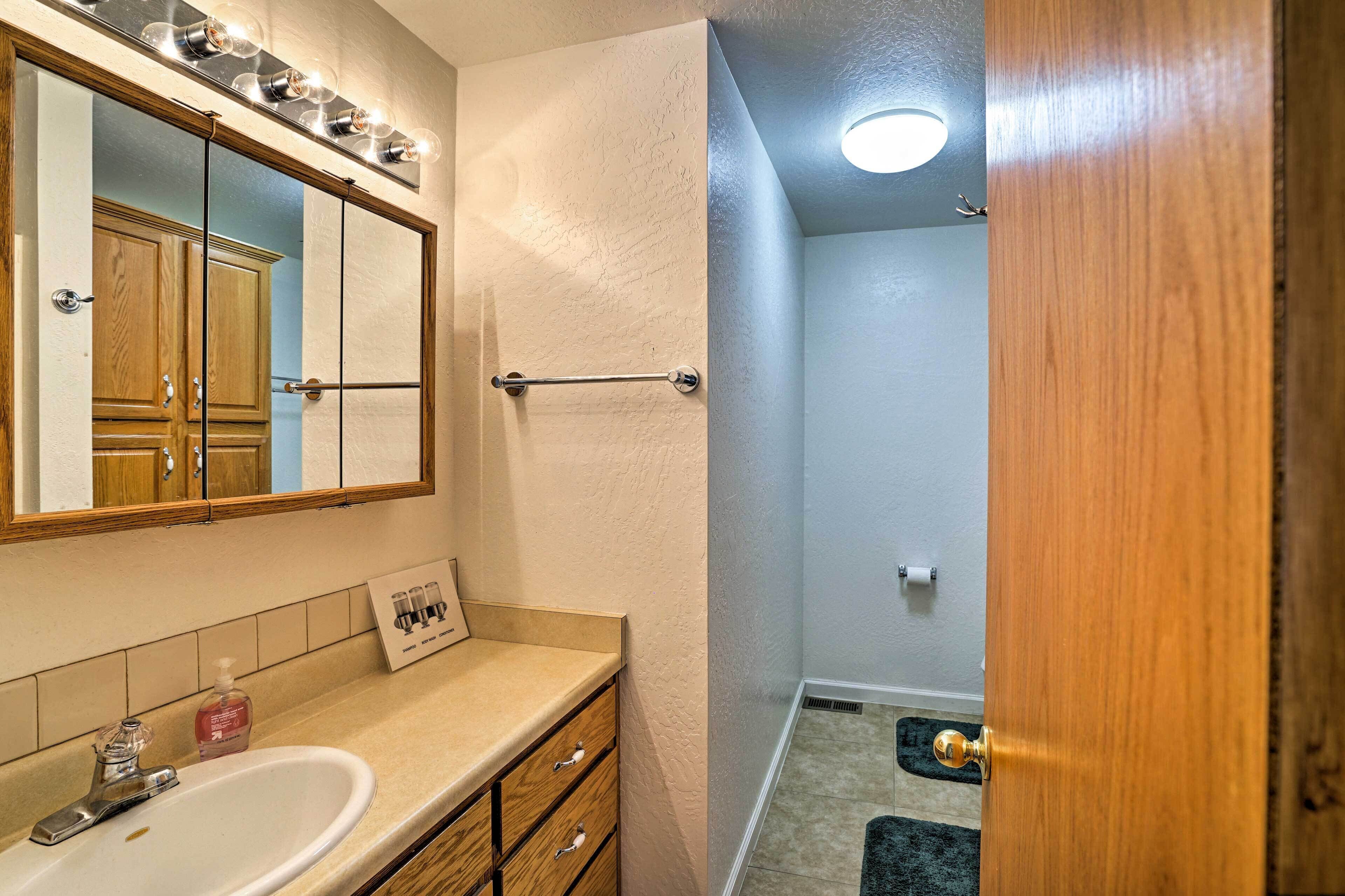 Une salle de bain avec un lavabo blanc, un meuble-lavabo en bois et un miroir avec éclairage intégré. Une porte donne accès à l’espace douche.