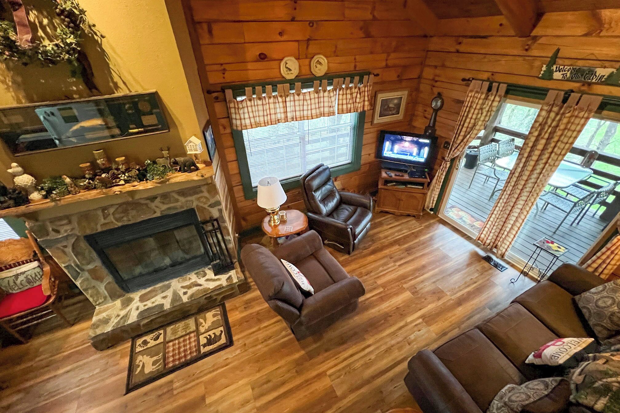 L’intérieur chaleureux d’un chalet avec une cheminée en pierre, des fauteuils en cuir, un téléviseur et une terrasse en bois visible à travers les fenêtres.