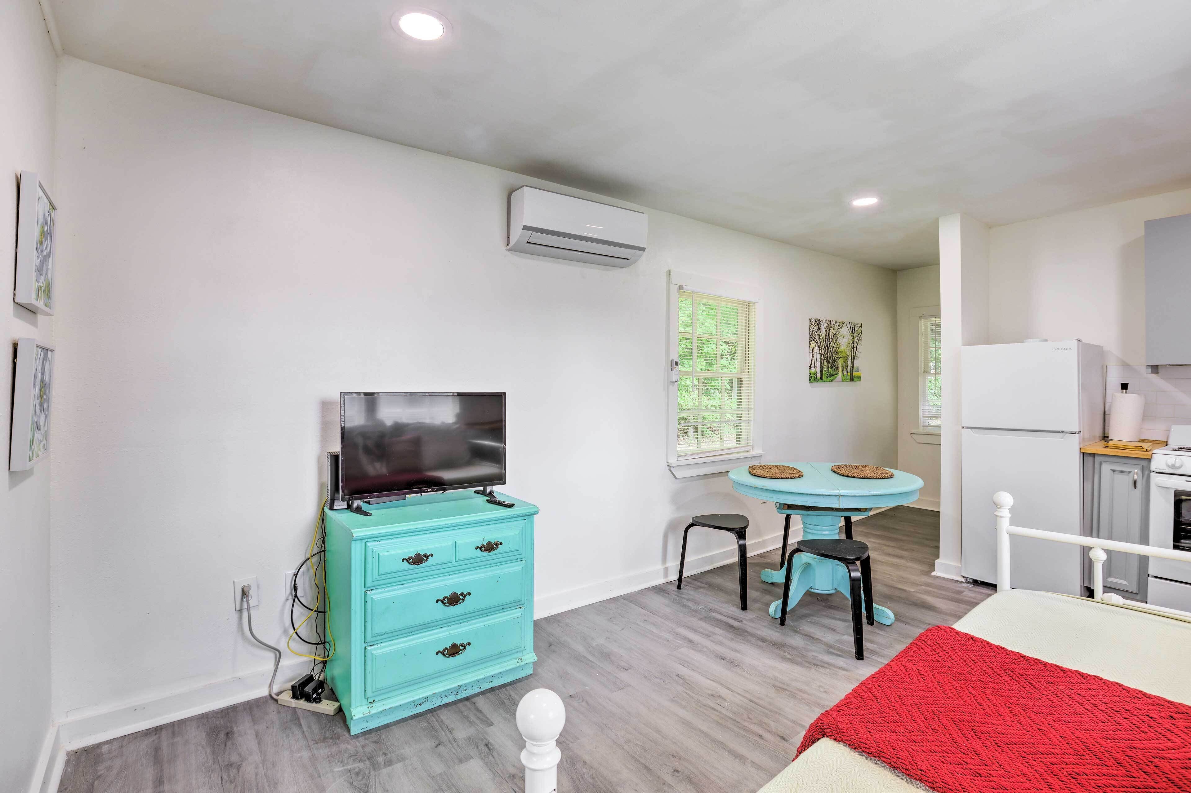 Une pièce de taille modeste, avec une commode de couleur turquoise, un téléviseur, une petite table ronde avec une chaise, un lit blanc avec une couverture rouge et un coin cuisine.