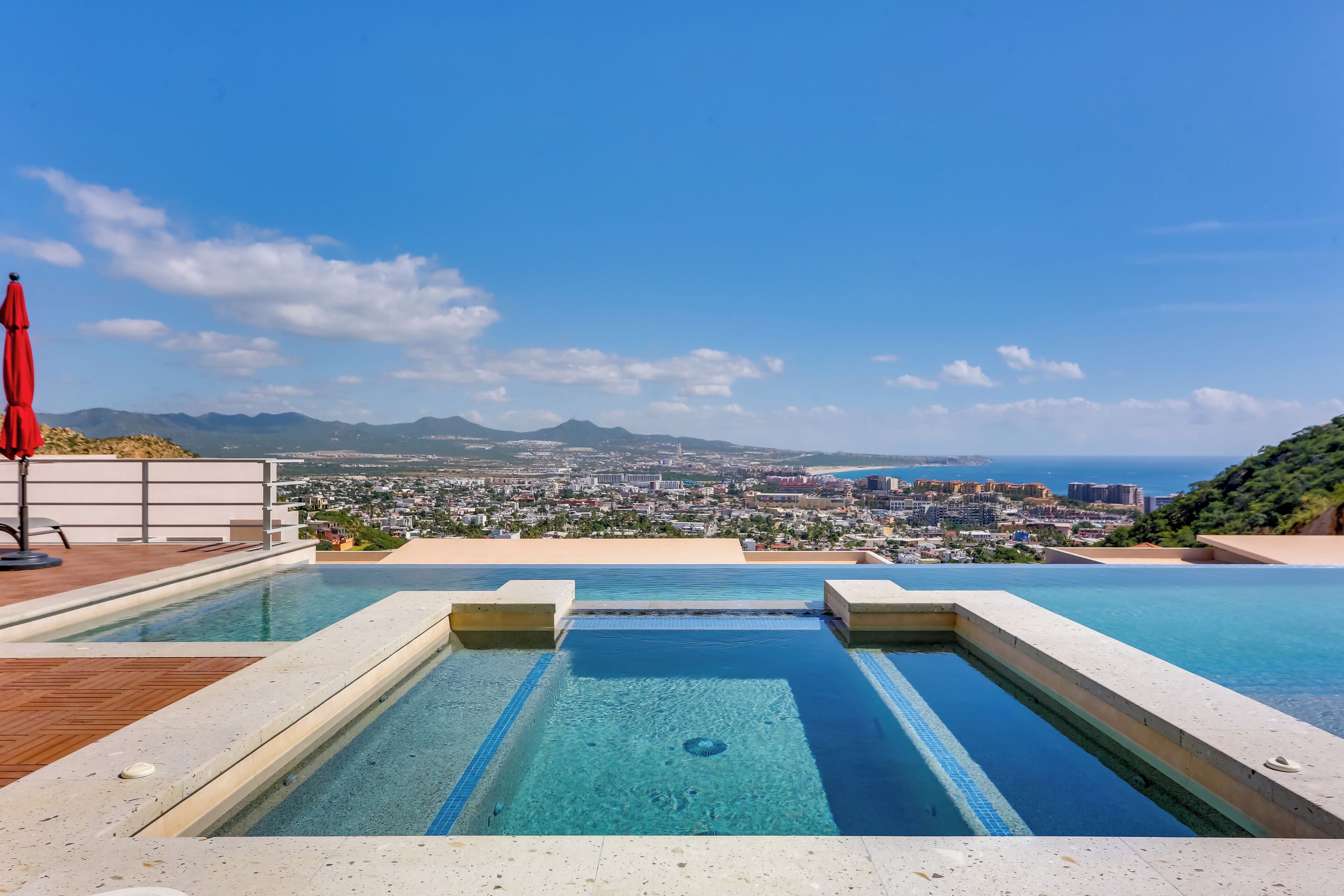 A rooftop infinity pool with a city view, a red umbrella, and a wooden deck.