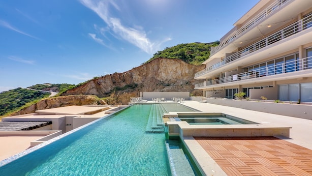 A modern hotel with a large infinity pool, outdoor seating, and a view of a cliff. - Cabo Pedregal Condo: Ocean View & 2 Infinity Pools (Cabo San Lucas)
