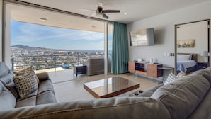 A modern living room with a large glass door leading to a balcony, a ceiling fan, a flat-screen TV, and a sofa with patterned cushions. - Cabo Pedregal Condo: Ocean View & 2 Infinity Pools (Cabo San Lucas)