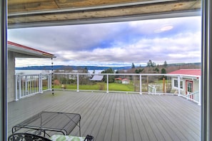 Una terraza de madera con una mesa y sillas de metal, vista a una casa con techo rojo y un cielo nublado.