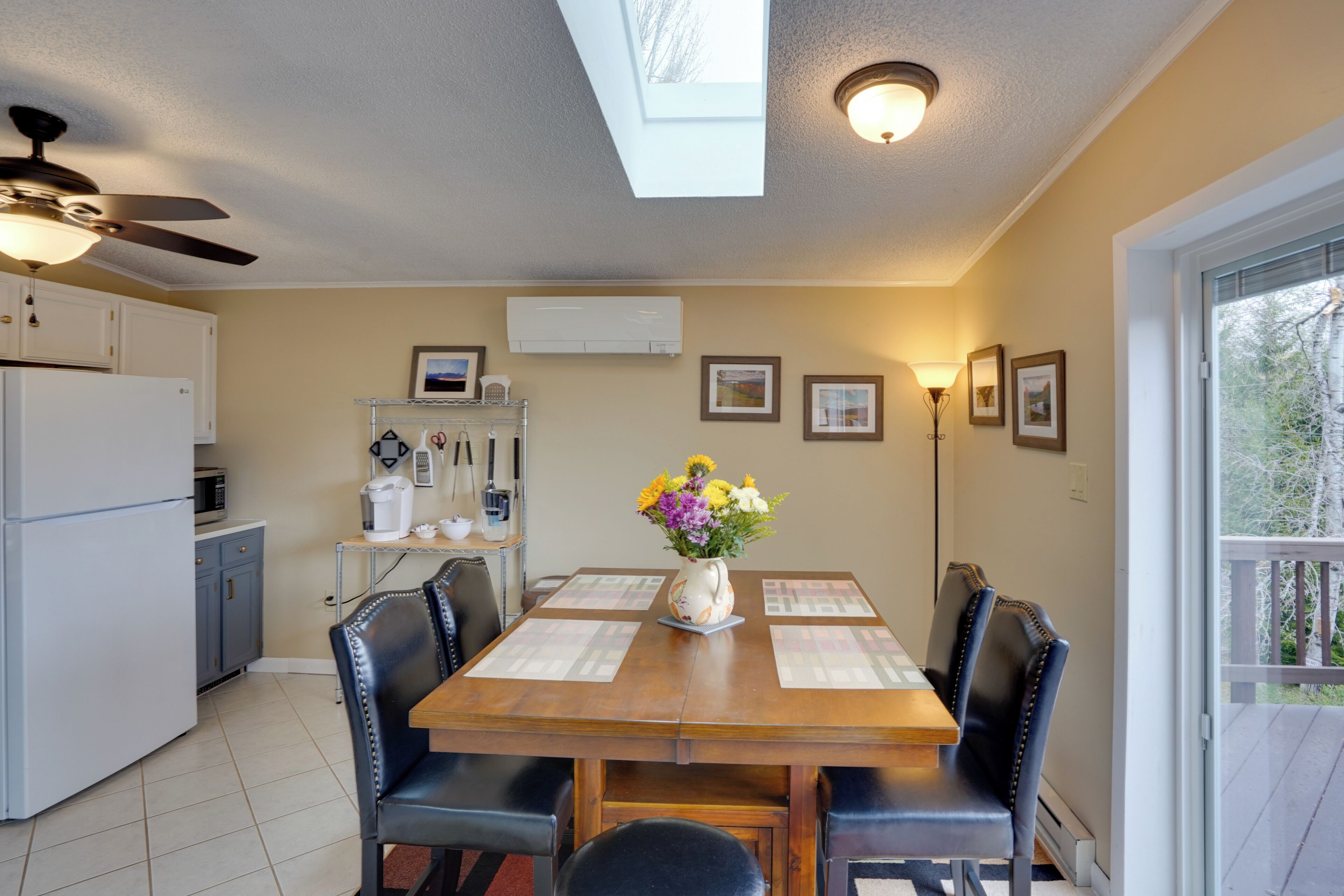 A dining area with a wooden table, black chairs, and a vase of flowers. There is a ceiling fan, a window with a view, and a kitchen area with cabinets and appliances.