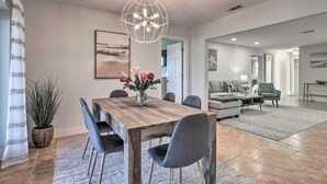 A spacious living area with a wooden dining table, grey chairs, a globe chandelier, and a large window with curtains. - Beach House w/ Pool: 3 Mi to Anna Maria Island! (Bradenton)