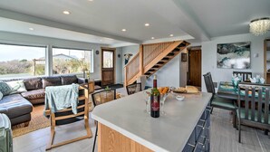 Amplia sala de estar con un ventanal grande, una escalera de madera, un comedor con mesa y sillas, y una isla de cocina con una botella de vino.