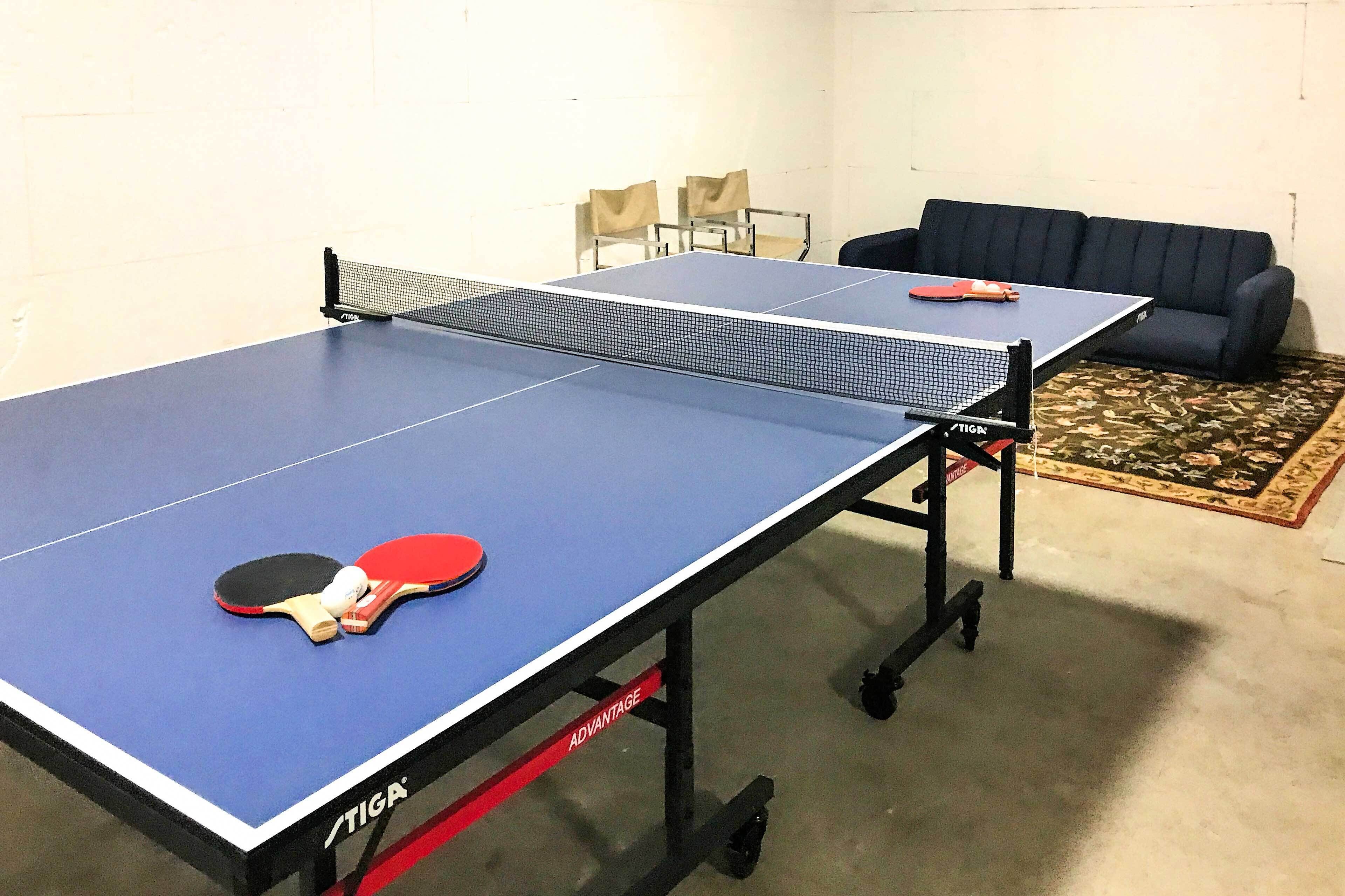 A ping pong table with a net, two chairs, and a sofa in a room with a carpeted floor.