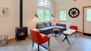 A living room with a fireplace, a sofa, a red chair, and a large window. - Modern Beaver Valley Cabin w/ Hot Tub & Games! (Leavenworth)