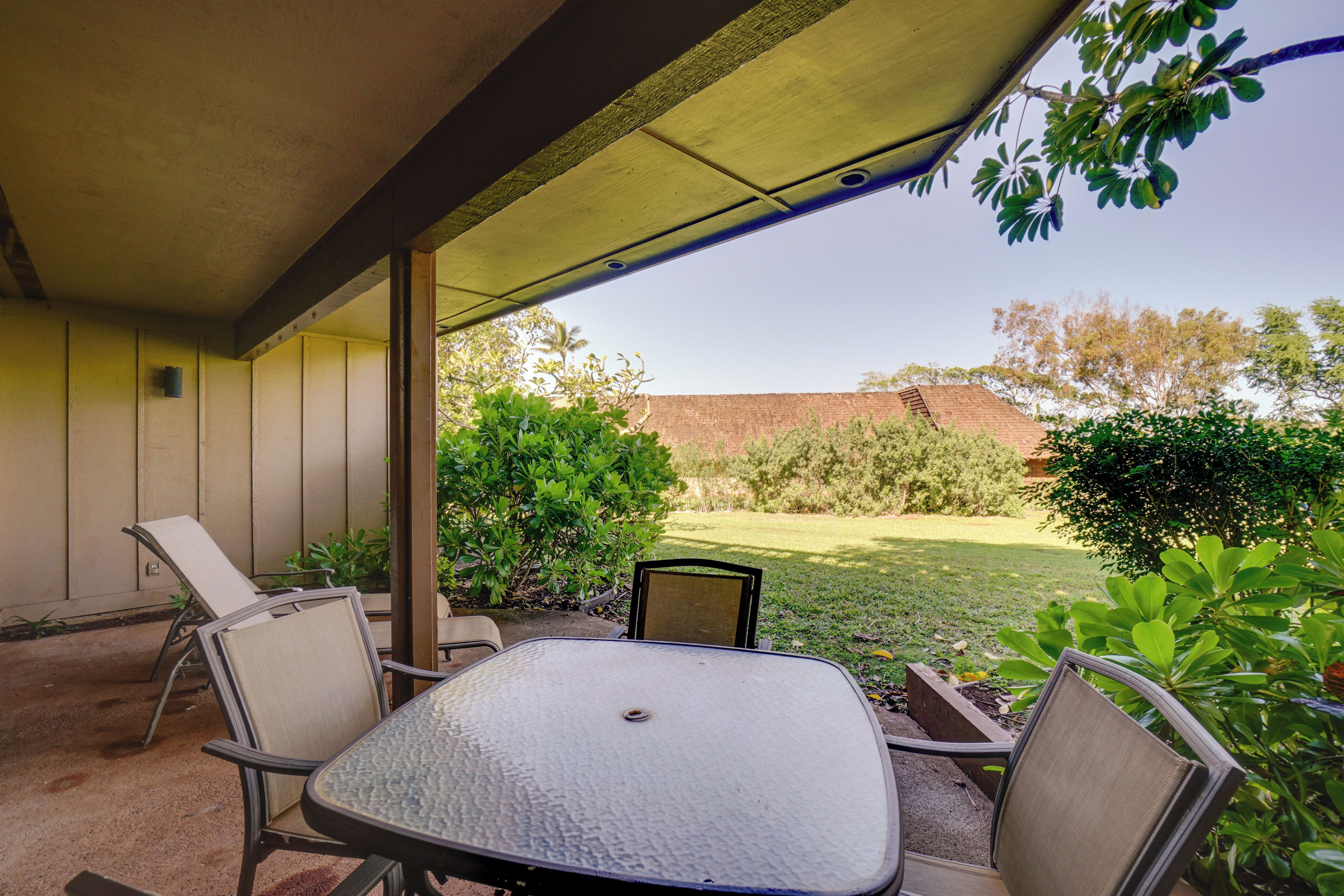 An outdoor patio with a table and chairs, a covered area, and a view of a grassy yard and trees.