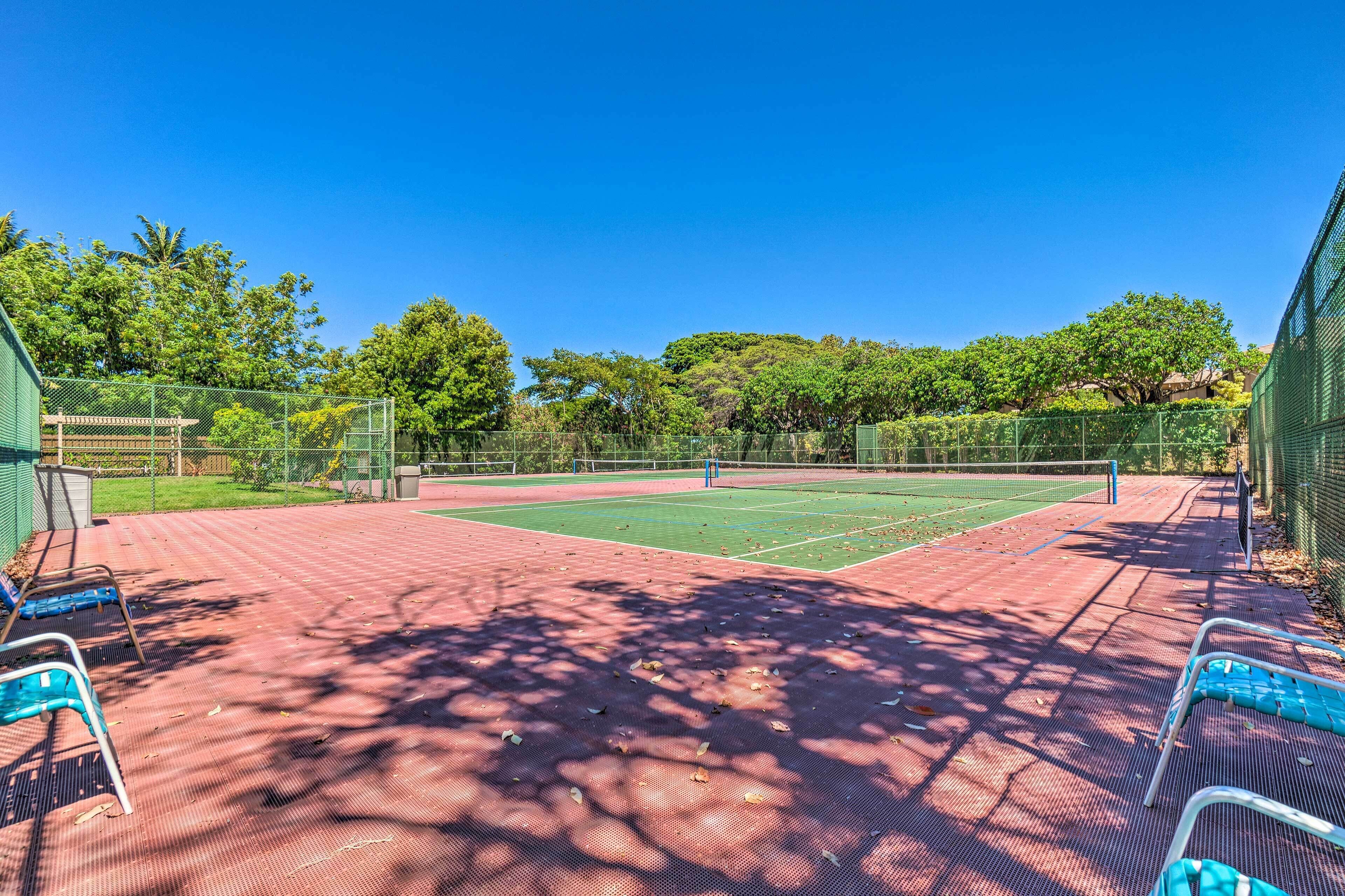 A tennis court with a red surface, surrounded by a green fence, and shaded by trees.