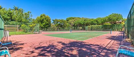 A tennis court with a red surface, surrounded by a green fence, and shaded by trees.