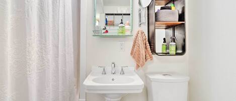 A bathroom with a white sink, two faucets, a toilet, a mirror, and a shelf with toiletries.