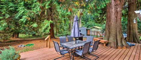 Een houten terras met een tafel en stoelen, omringd door bomen en een kleine schuur.