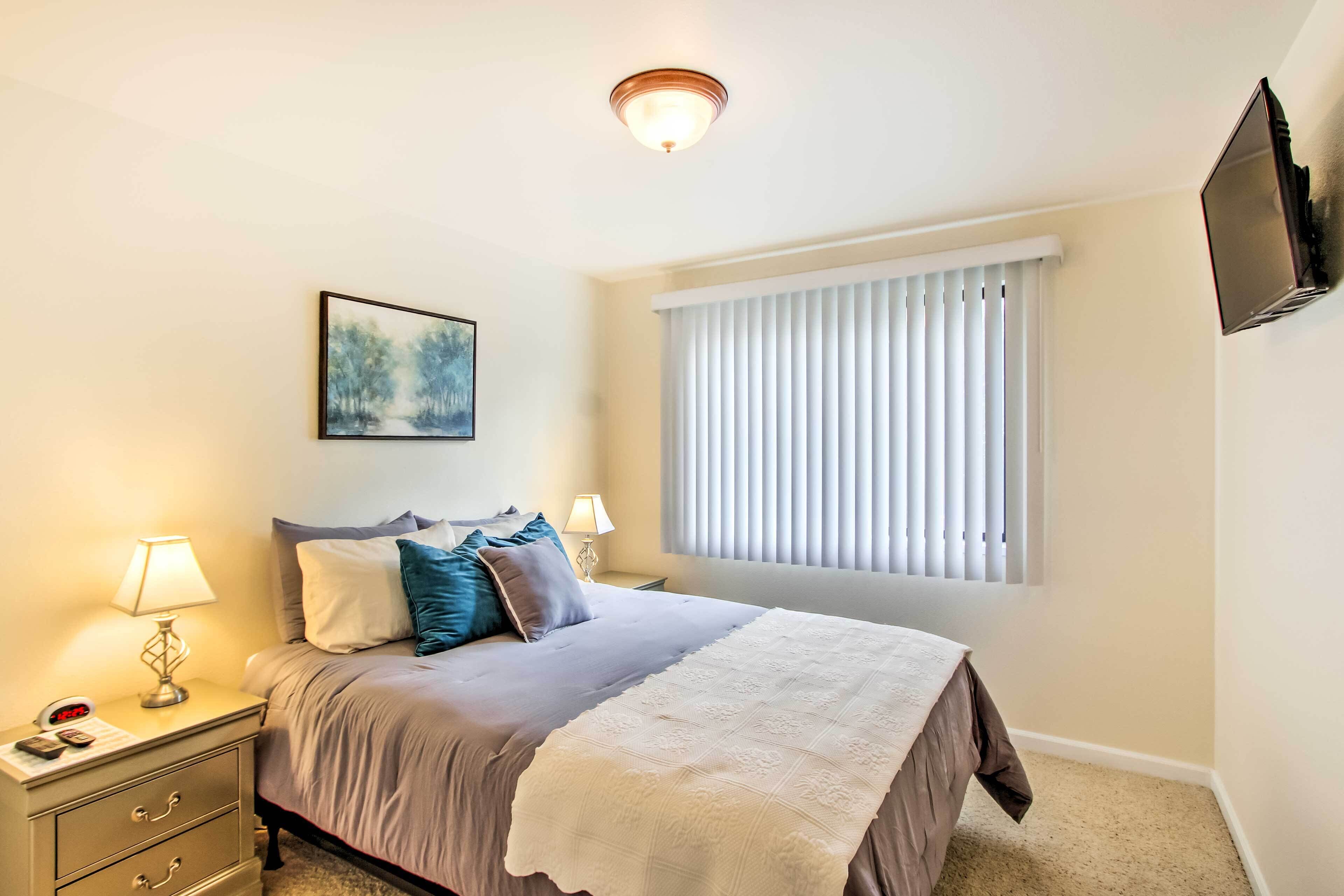 A neatly arranged bedroom with a bed, bedside lamps, a nightstand, a framed picture, a window with blinds, and a mounted TV.