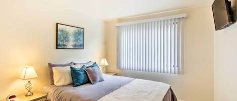 A neatly arranged bedroom with a bed, bedside lamps, a nightstand, a framed picture, a window with blinds, and a mounted TV.