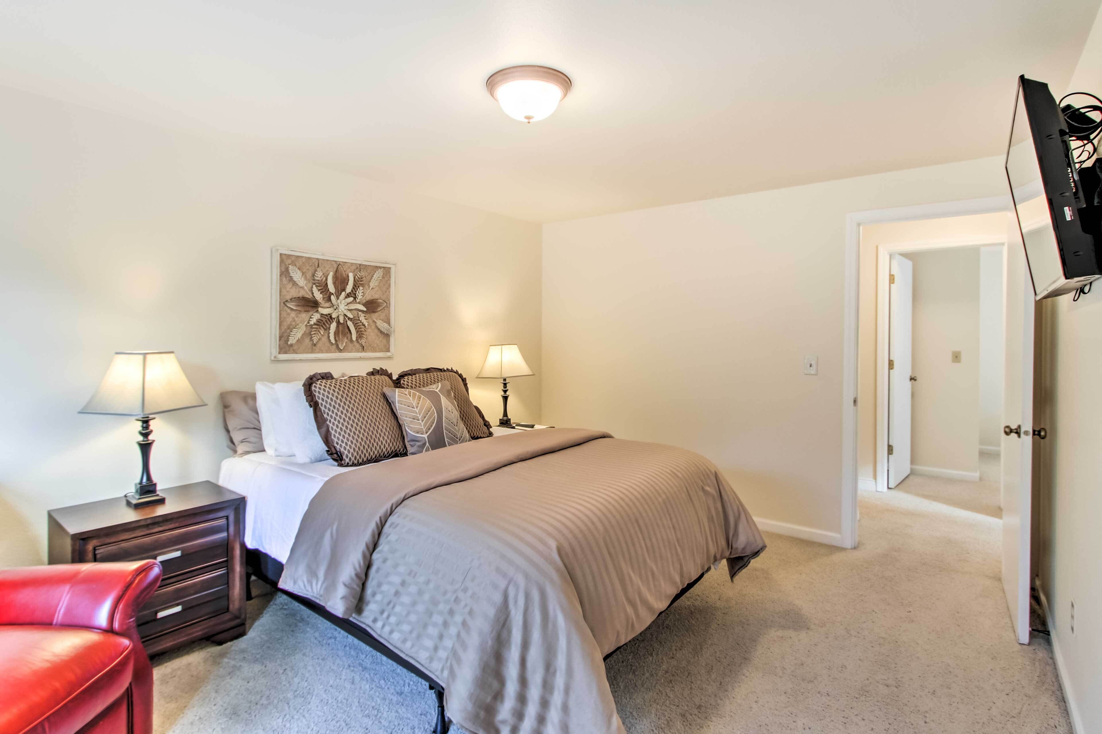 A bedroom with a bed, two bedside tables with lamps, a red chair, a decorative wall piece, and a door leading to another room.
