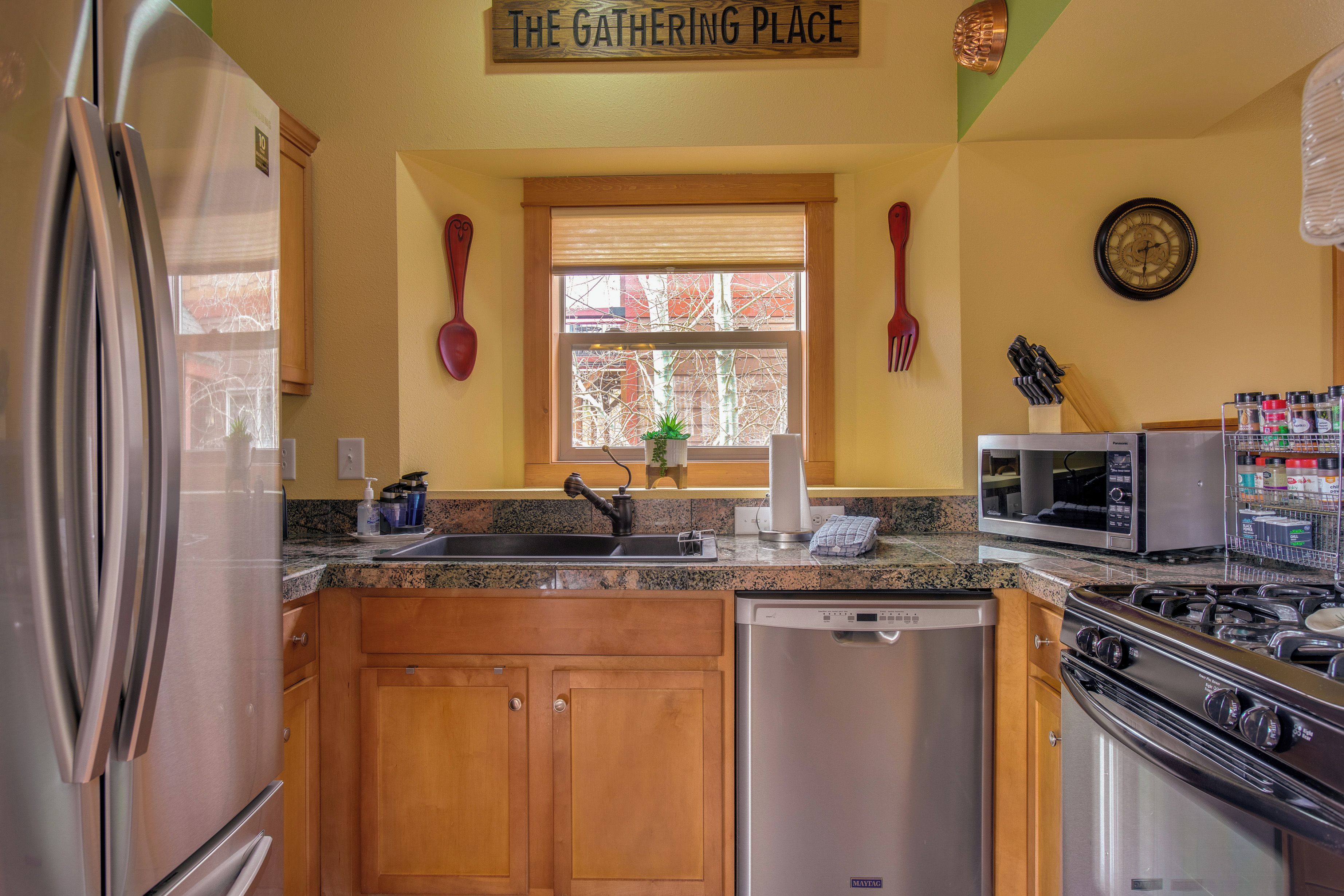 A kitchen with a stainless steel refrigerator, a dishwasher, a microwave, and a stove. There is a window with blinds, a clock on the wall, and a sign above the window that reads "The Gathering Place".