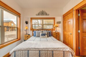 A bedroom with a large bed, wooden headboard, two bedside tables with lamps, a window with a view of buildings, and a wooden door. - Riverfront Cabin, 4 Mi to Granby Ranch (Granby)