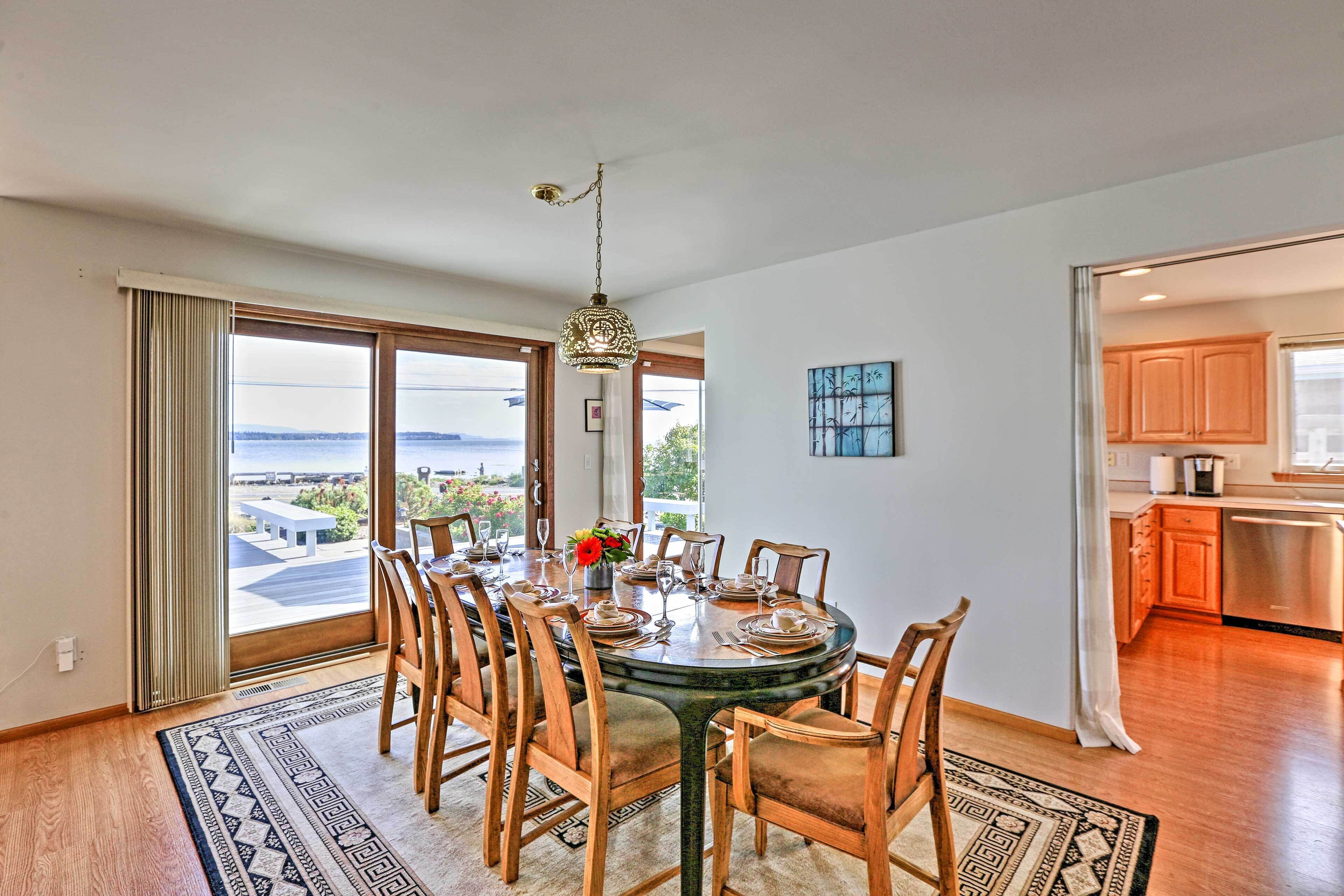 A dining area with a round table set for six, wooden chairs, a hanging light fixture, and a view through large windows.