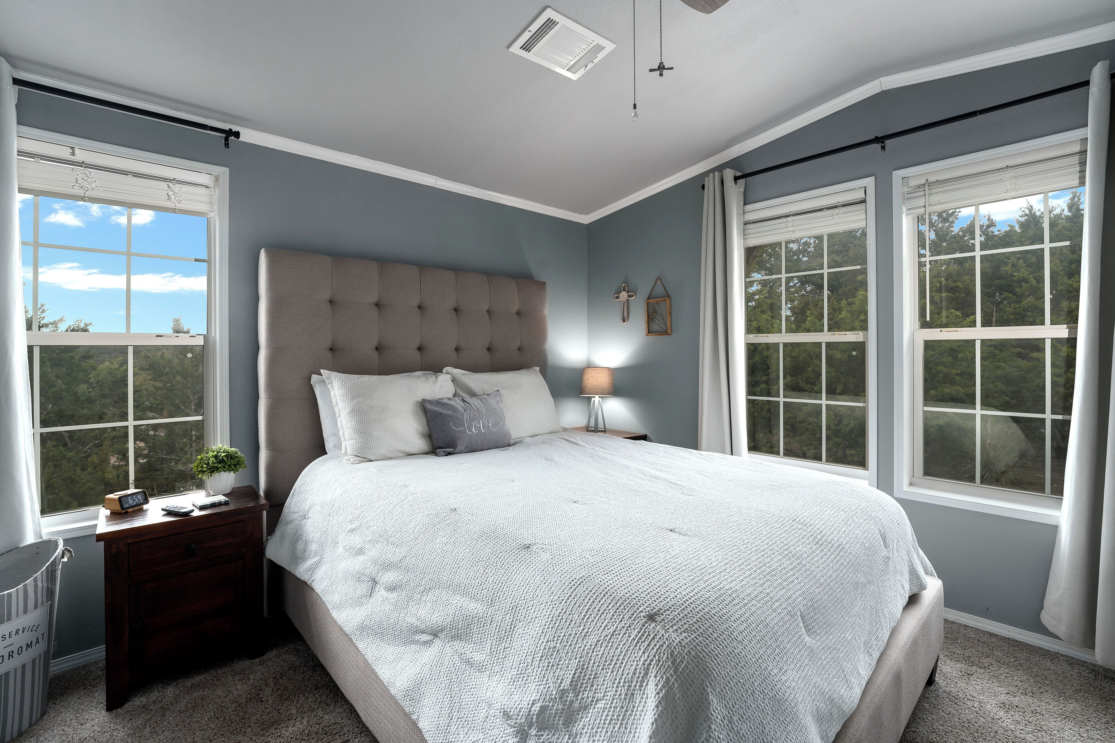 A bedroom with a large bed, a nightstand, a lamp, and a view of trees through the windows.