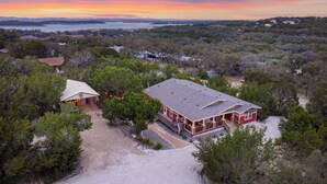 A large house with a spacious deck and a covered area, surrounded by dense trees and a view of the ocean at sunset. - Hill Country Gem: Sunset Deck, Hot Tub & Fire Pit (Canyon Lake)