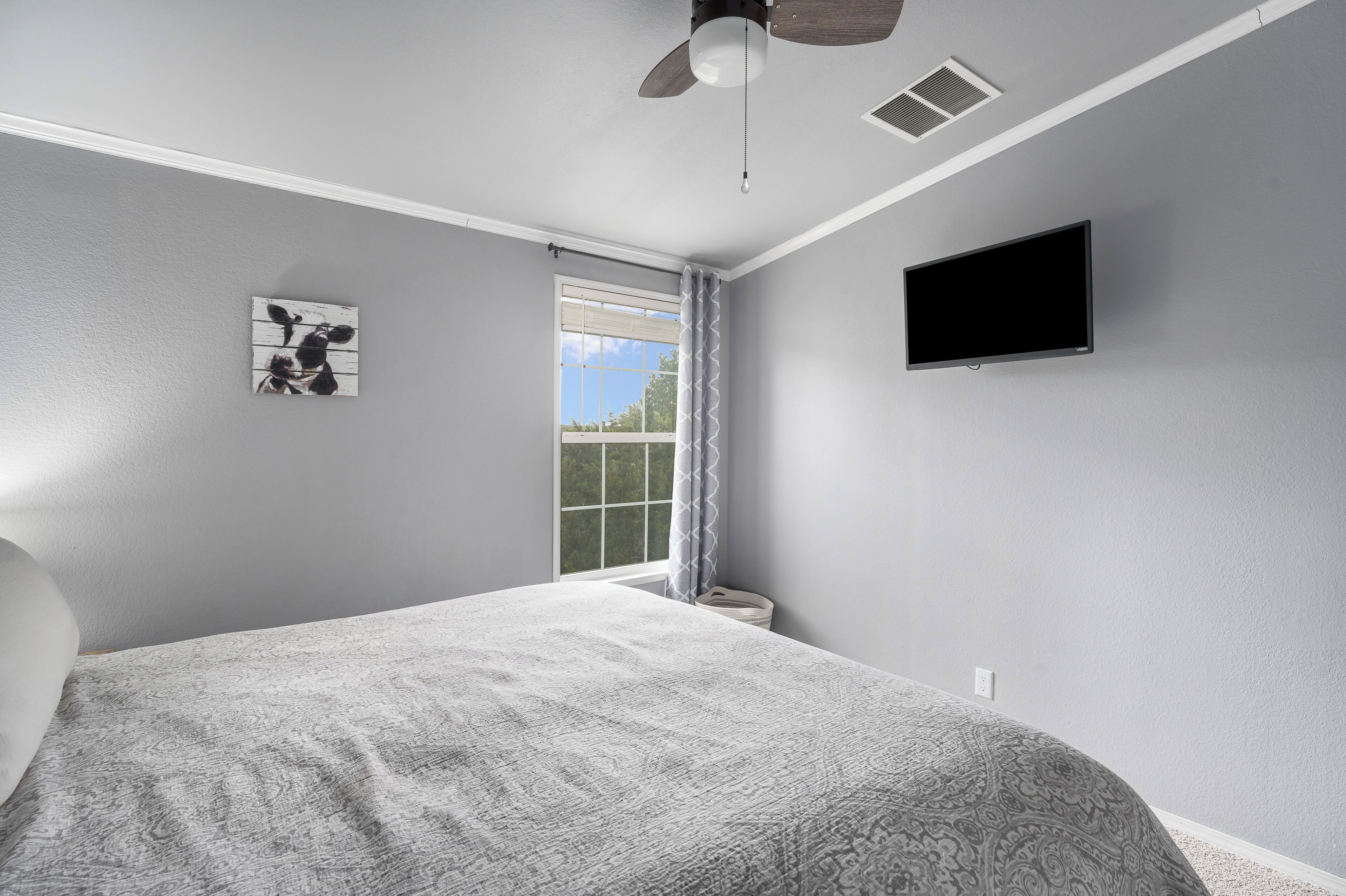 Ein Schlafzimmer mit Bett, Deckenventilator, Fenster mit Vorhängen und einem Wand-Fernseher.