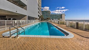 A swimming pool with a 6 ft depth marker, surrounded by a brick deck and white railings, with a building in the background.